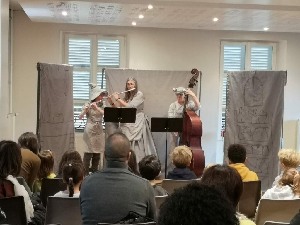 Les trois comédiennes, deux elfes et un crapaud, sont sur scène et jouent de la musique,avec deux flûtes traversières et une contrebasse. Les décors et les costumes sont gris.